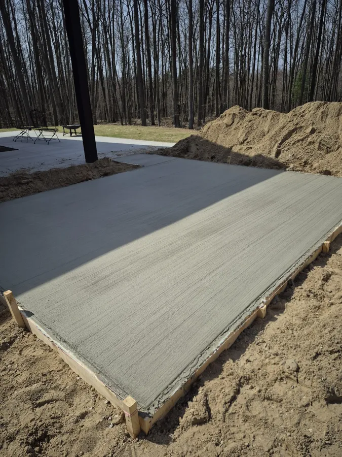 Long broom-finished concrete slab poured beside an asphalt drive, forms still staked, woods behind