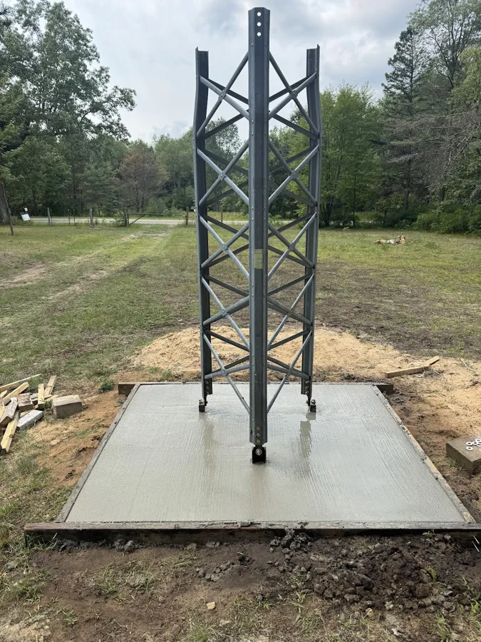 Square concrete pad poured as the base for a steel lattice tower in a grassy yard